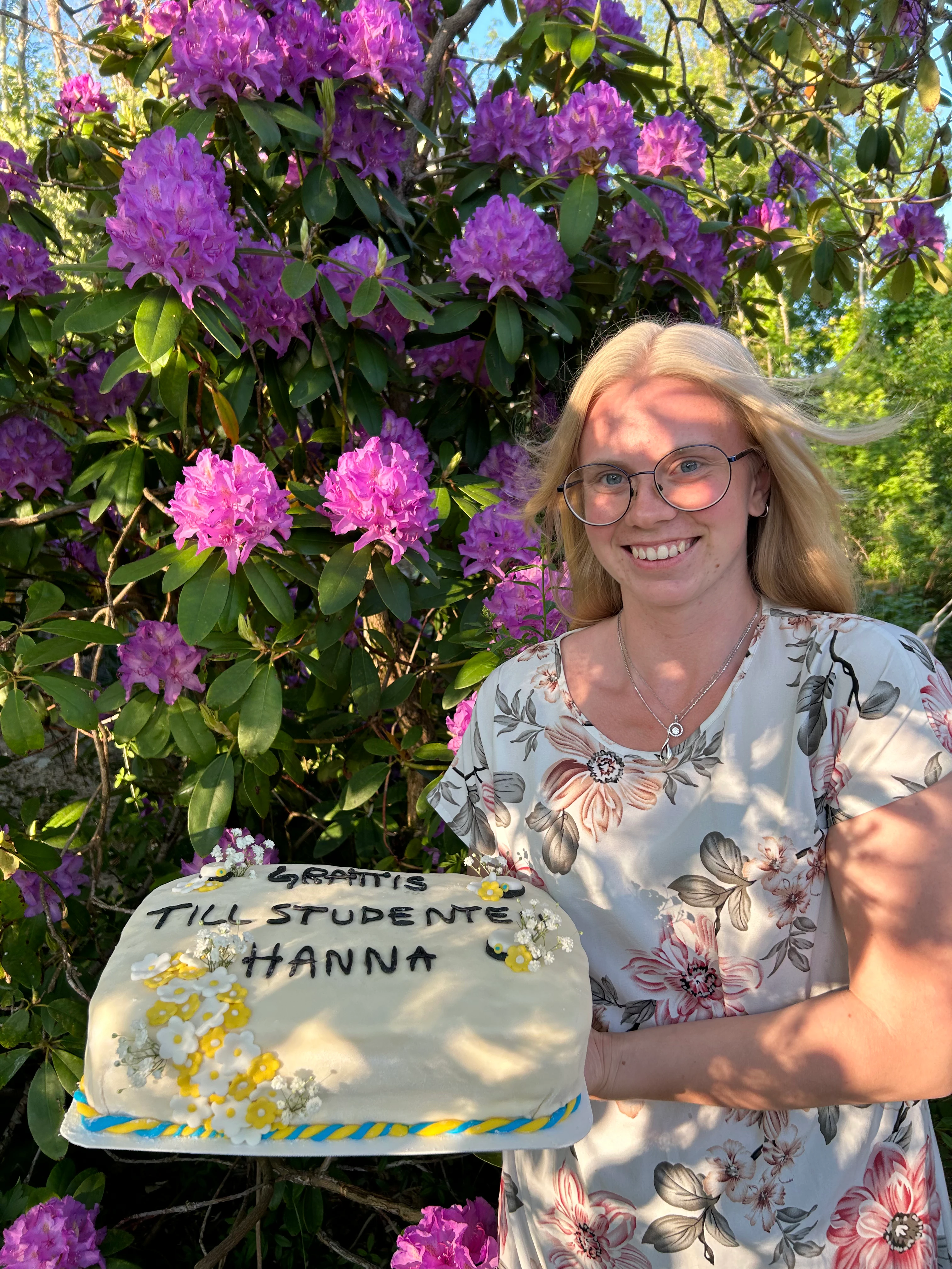 Josefine står vid en rhododensdron och visar upp en studenttårta